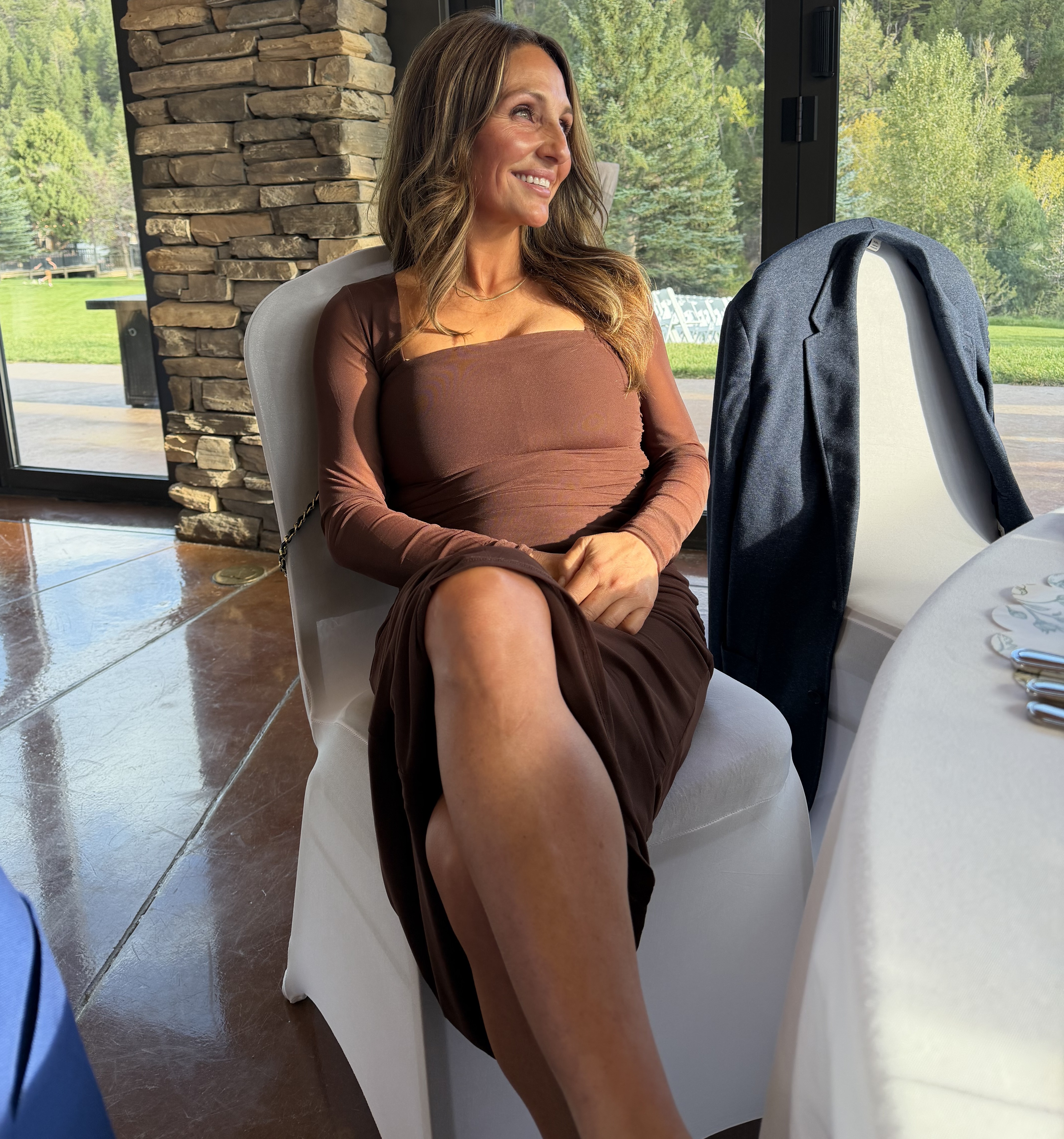 Smiling woman in a brown long-sleeve dress at a mountain wedding venue, showing a warm, natural-looking bronze spray tan